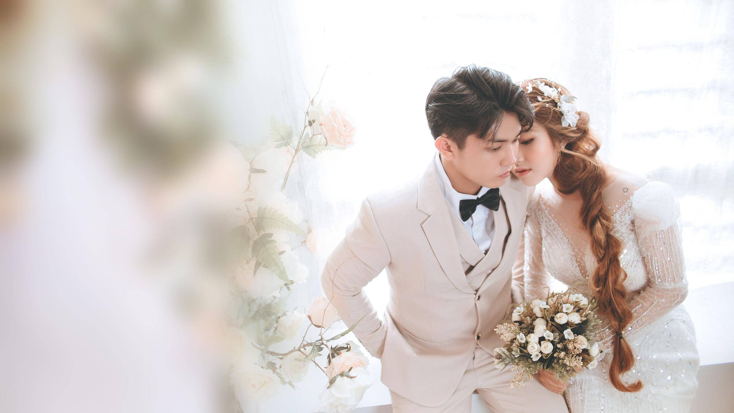 A bride holding a small bouquet leans against a groom in a cream suit in a bright, airy room with white roses