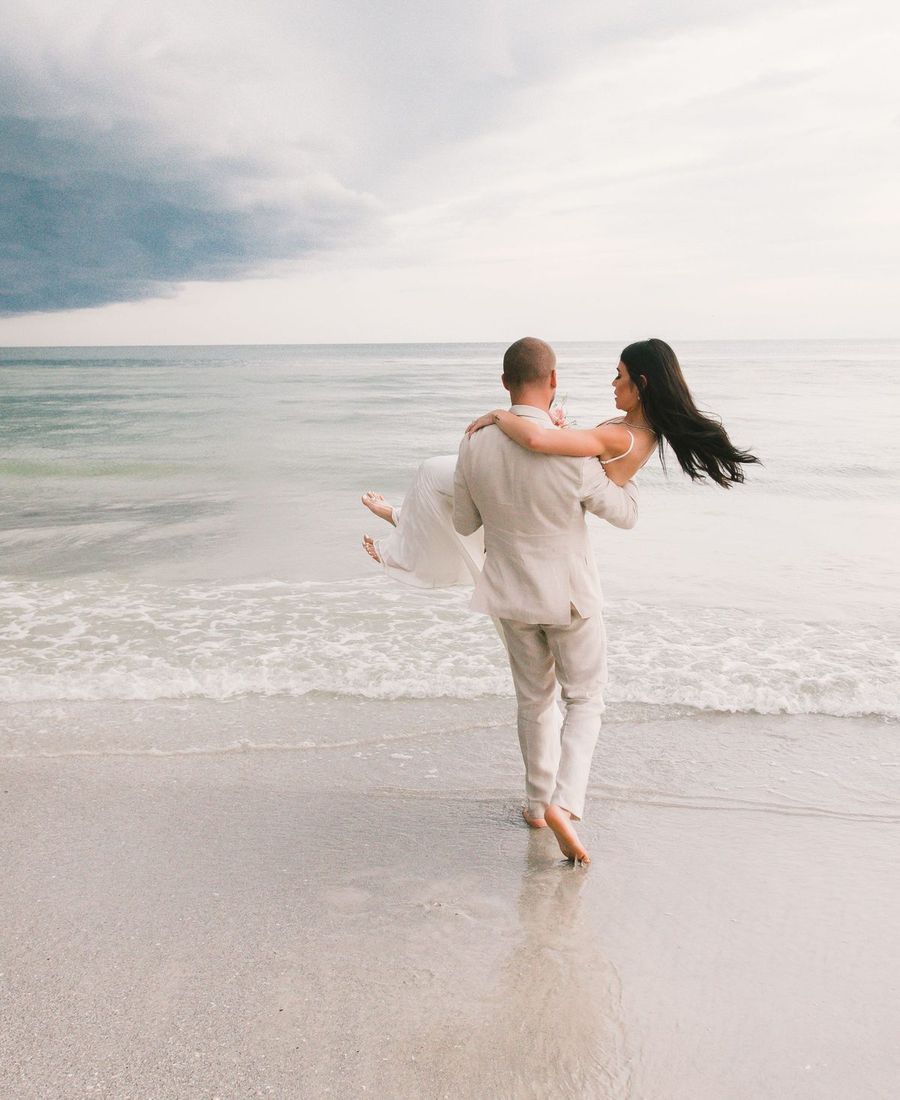 A groom in a linen suit carries his bride into the shallow surf on a pale, quiet beach, seen from behind