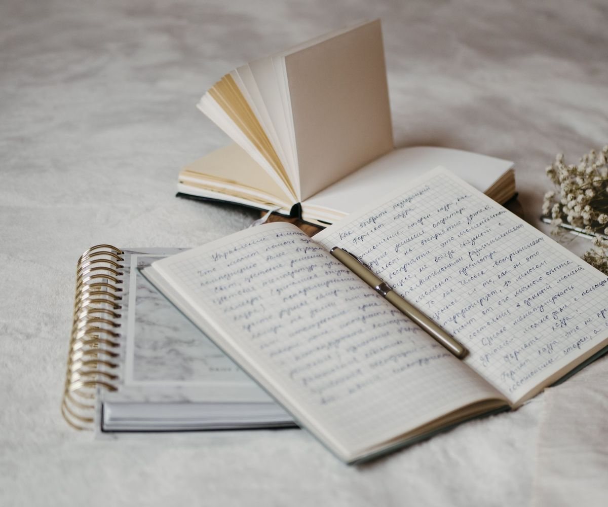 Open notebooks with handwritten notes, a gold spiral planner, a pen and a sprig of dried baby's breath on a soft white blanket